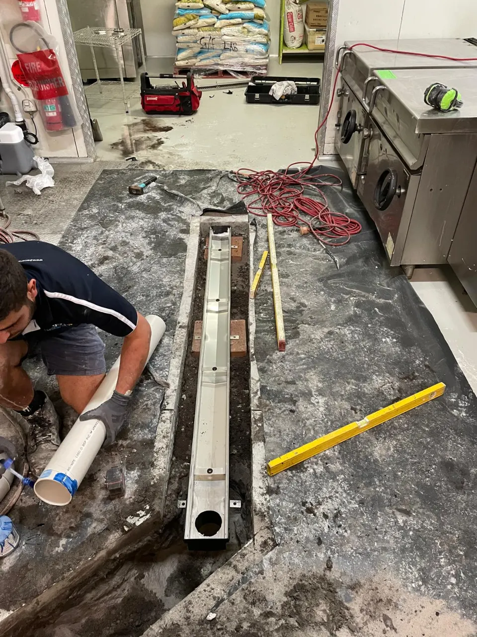 Pacer plumber fitting PVC pipe to a stainless steel channel drain set into a cut-out in a commercial kitchen floor