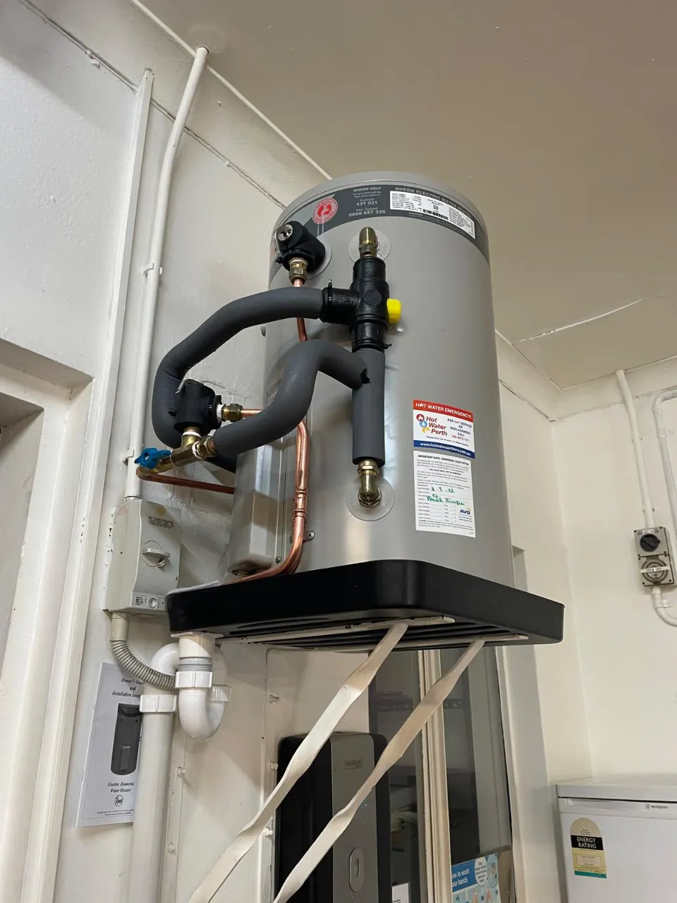 Storage hot water heater on a black bracket with insulated pipework and a white waste trap underneath