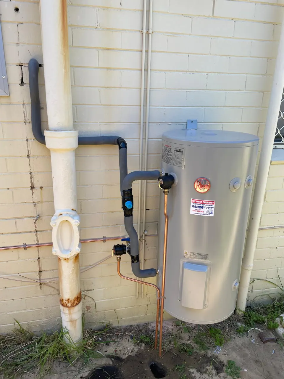 Rheem electric storage hot water system against a brick wall beside a downpipe, with insulated pipework and a copper relief line