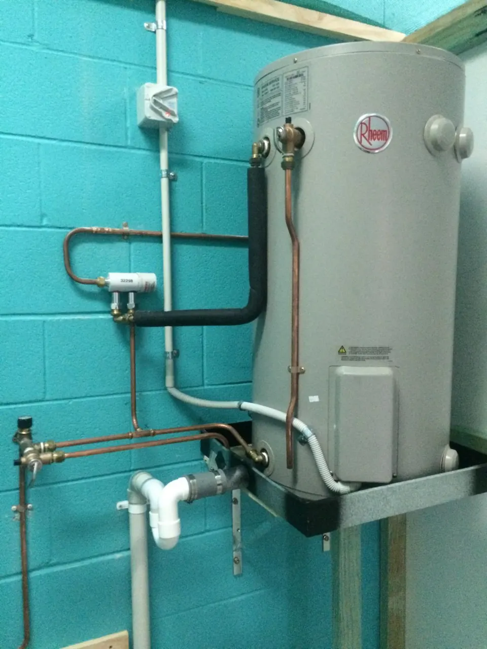 Rheem electric storage hot water heater on a steel bracket against a teal block wall, with copper pipework and an isolator switch