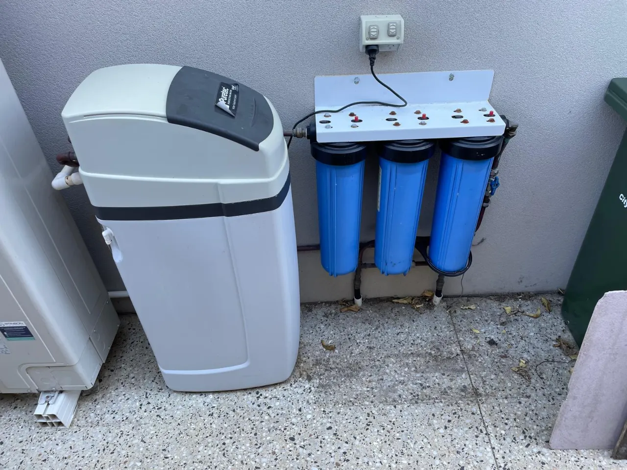 Water softener and a three-stage filter set with blue housings mounted on a wall outside a home