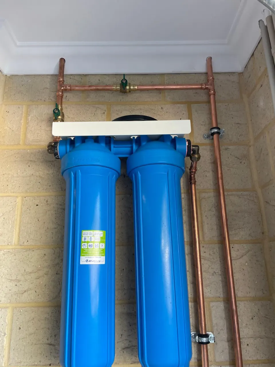 Two-stage blue whole-house water filter mounted on a brick wall with copper pipework and isolation valves