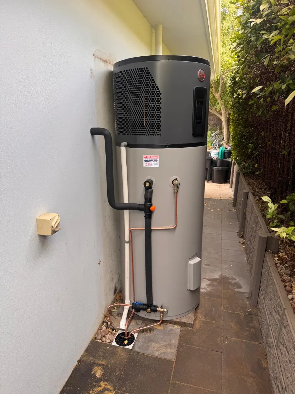 Heat pump hot water system on a rendered wall in a paved side passage, with insulated pipework and a copper drain line