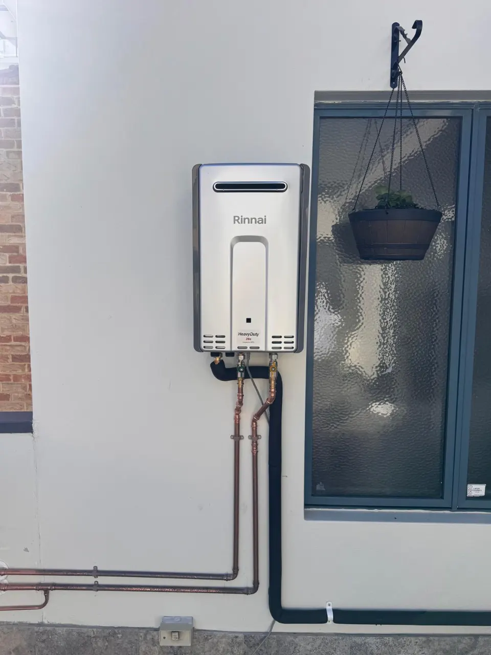 Rinnai continuous flow gas hot water unit mounted on a rendered wall beside a window, with copper and black pipework
